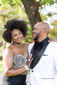  Close-up of a joyful couple at an outdoor wedding, with the woman in a glamorous black and white beaded strapless dress and the man in a dapper white tuxedo jacket and black bow tie. They are smiling warmly at each other, showcasing their happiness and love amidst a backdrop of lush greenery. Perfect for showcasing a sophisticated and elegant wedding style.